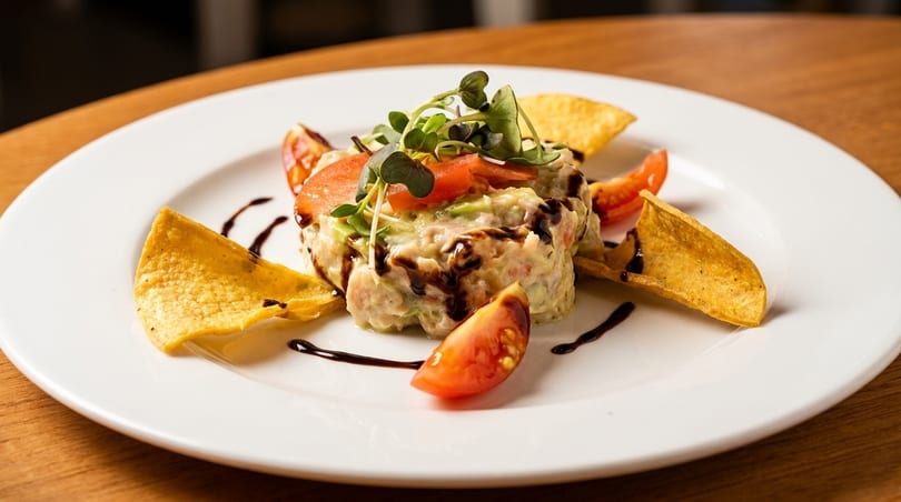 A white plate with a tuna and avocado tartare, garnished with tomatoes, microgreens, and tortilla chips.