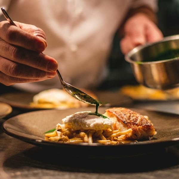Chef plating a dish with fish, pasta, and a poached egg, drizzling green sauce.