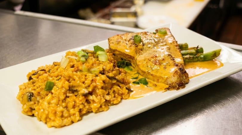 A white plate holds a salmon fillet with creamy sauce, risotto, and asparagus spears.